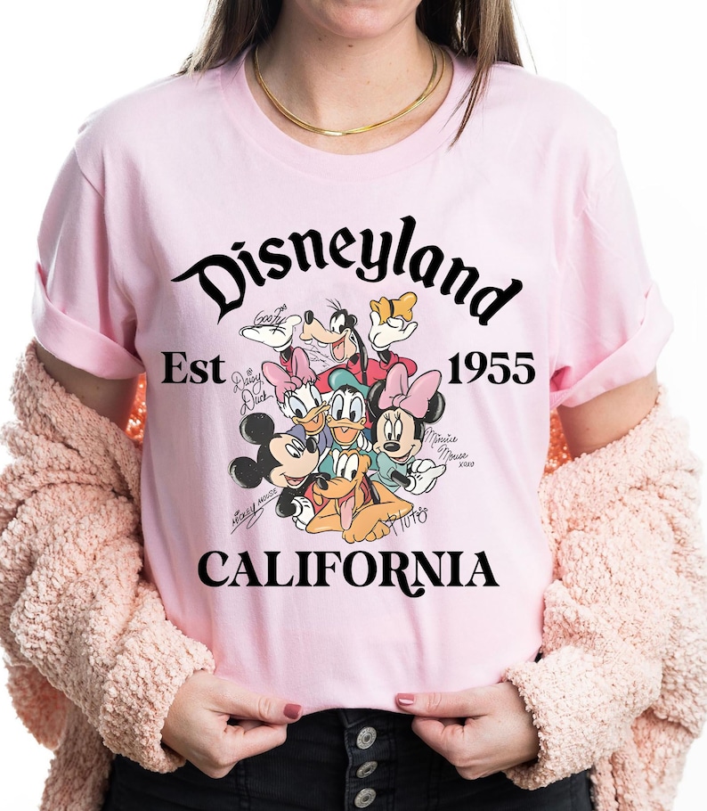May include: A light pink t-shirt featuring the Disneyland logo with the text "Est 1955 CALIFORNIA". The shirt has a graphic of Disney characters including Mickey Mouse, Minnie Mouse, Donald Duck, and Goofy.