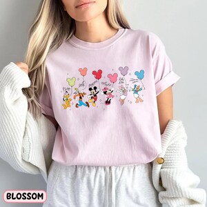 K&ouml;nnte beinhalten: Hellrosa T-Shirt mit einer Grafik von Disney-Figuren, die Luftballons halten. Die Figuren sind Pluto, Goofy, Mickey Mouse, Minnie Mouse, Daisy Duck und Donald Duck. Das Wort "Blossom" ist unten aufgedruckt.