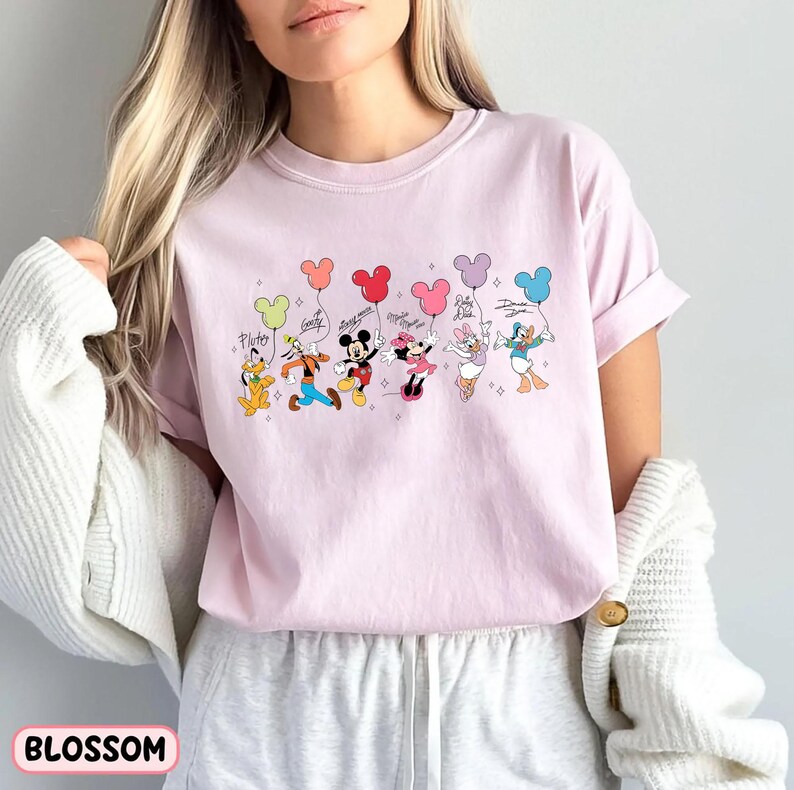 K&ouml;nnte beinhalten: Hellrosa T-Shirt mit einer Grafik von Disney-Figuren, die Luftballons halten. Die Figuren sind Pluto, Goofy, Mickey Mouse, Minnie Mouse, Daisy Duck und Donald Duck. Das Wort "Blossom" ist unten aufgedruckt.