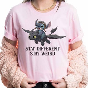 May include: Pale pink t-shirt with a cartoon image of Stitch riding a dragon. Stitch wears a pilot's hat and goggles. The text "STAY DIFFERENT STAY WEIRD" is printed beneath the graphic in a bold font.