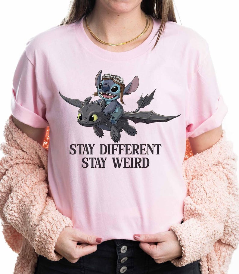 May include: Pale pink t-shirt with a cartoon image of Stitch riding a dragon. Stitch wears a pilot's hat and goggles. The text "STAY DIFFERENT STAY WEIRD" is printed beneath the graphic in a bold font.
