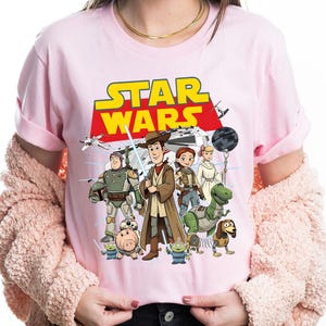 May include: Pale pink t-shirt with a graphic of Star Wars characters, including Woody and Buzz Lightyear. The words "STAR WARS" are in yellow and red. The shirt has a crew neck.