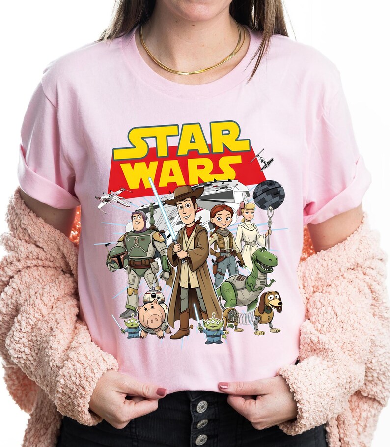 May include: Pale pink t-shirt with a graphic of Star Wars characters, including Woody and Buzz Lightyear. The words "STAR WARS" are in yellow and red. The shirt has a crew neck.