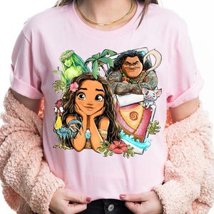 May include: Pink t-shirt featuring a colorful graphic of characters from a popular animated movie. The design includes a central female character with long brown hair, surrounded by other characters and tropical elements.