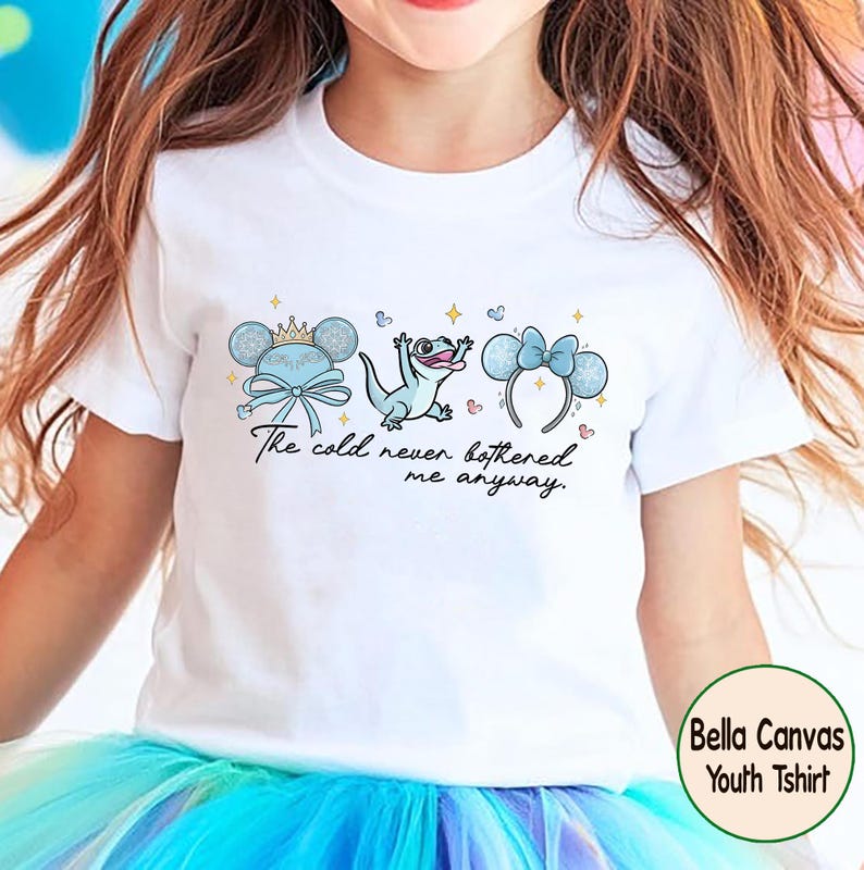 Op de afbeelding: Wit kinder-T-shirt met een grillig ontwerp met een kroon, strik en muisoren. In het midden staat een lichtblauwe hagedis met de tekst "The cold never bothered me anyway." Het T-shirt heeft een ronde hals en korte mouwen.