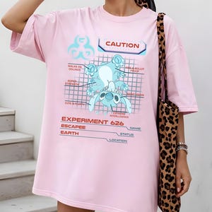 May include: A light pink t-shirt featuring a blue cartoon character design with the text "CAUTION" and "EXPERIMENT 626". The graphic includes the words "WALKS ON CEILINGS", "FIRE & BULLET PROOF", "SUPER STRENGTH", "CUTE & FLUFFY", "NIGHT VISION", and "EXTREME INTERLLIGENCE".