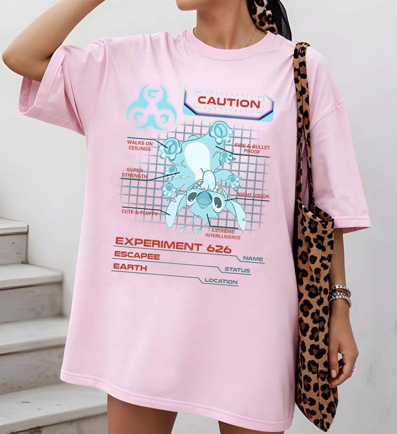 May include: A light pink t-shirt featuring a blue cartoon character design with the text "CAUTION" and "EXPERIMENT 626". The graphic includes the words "WALKS ON CEILINGS", "FIRE & BULLET PROOF", "SUPER STRENGTH", "CUTE & FLUFFY", "NIGHT VISION", and "EXTREME INTERLLIGENCE".