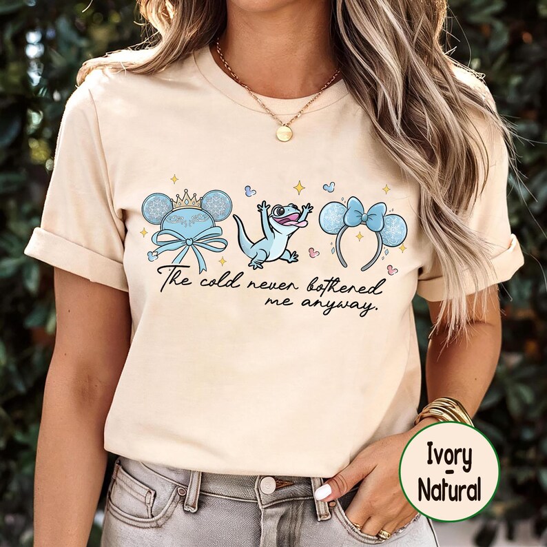 Op de afbeelding: Cr&egrave;mekleurig T-shirt met een afbeelding van een cartoon hagedis en twee sets muizenoren met strikken en kronen. De tekst "The cold never bothered me anyway" staat onder de afbeelding.