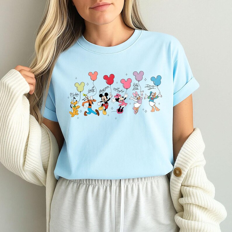 K&ouml;nnte beinhalten: Hellblaues T-Shirt mit einer Grafik von Disney-Figuren, die Mickey Mouse-f&ouml;rmige Ballons halten. Die Figuren sind Pluto, Goofy, Mickey, Minnie, Daisy und Donald Duck. Die Namen der Figuren sind dar&uuml;ber geschrieben.