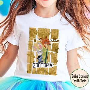 May include: White youth t-shirt featuring characters from the Disney movie Zootopia. The design includes Nick Wilde, Judy Hopps, and other characters in a gold-toned collage. The word "ZOOTOPIA" is printed in black. The shirt is a Bella Canvas youth t-shirt.