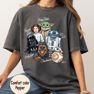 May include: Dark grey t-shirt featuring Star Wars characters: Darth Vader, Princess Leia, Baby Yoda, Chewbacca, R2-D2, and BB-8. The shirt has the text "Comfort color Pepper".