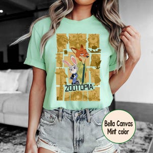 May include: A mint green t-shirt featuring a graphic from the Disney movie Zootopia. The design includes characters Judy Hopps and Nick Wilde, along with other characters in a grid pattern. The text "ZOOTOPIA" is below the characters.