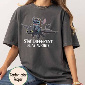 May include: Dark grey t-shirt with a cartoon image of Stitch riding a dragon, and the words "Stay Different Stay Weird". The shirt is a comfort colour pepper.