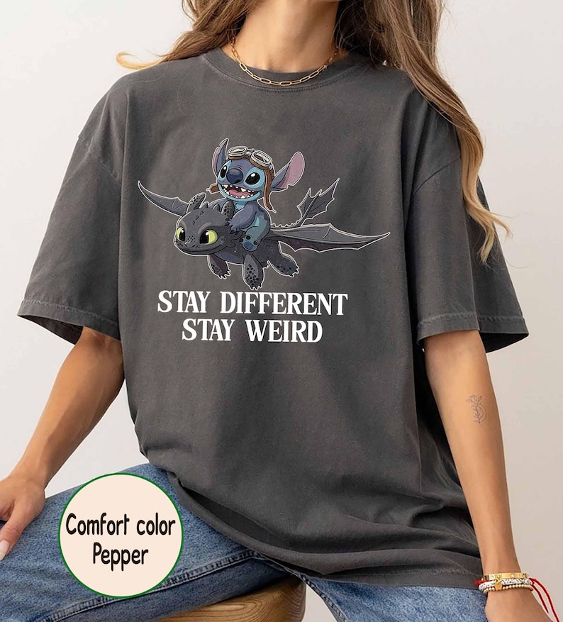 May include: Dark grey t-shirt with a cartoon image of Stitch riding a dragon, and the words "Stay Different Stay Weird". The shirt is a comfort colour pepper.