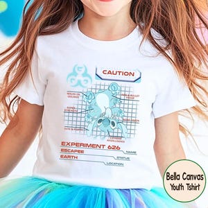 May include: White Bella Canvas youth t-shirt featuring a blue cartoon character design. The design includes the text "CAUTION" and "EXPERIMENT 626", along with other descriptive labels. The shirt is worn by a child.