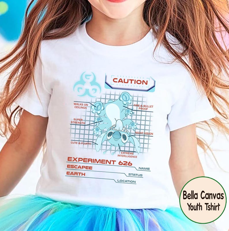 May include: White Bella Canvas youth t-shirt featuring a blue cartoon character design. The design includes the text "CAUTION" and "EXPERIMENT 626", along with other descriptive labels. The shirt is worn by a child.