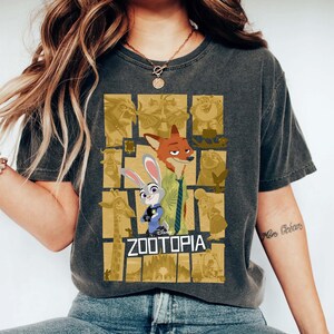 May include: Dark gray t-shirt featuring the word "ZOOTOPIA" with cartoon characters Nick Wilde and Judy Hopps. The shirt has a distressed look and a collage of Zootopia characters in the background.