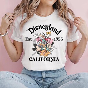 May include: White t-shirt featuring the Disneyland logo with the text "Est 1955 CALIFORNIA" in black script. The shirt showcases a colorful graphic of Disney characters including Mickey Mouse, Minnie Mouse, and Donald Duck.