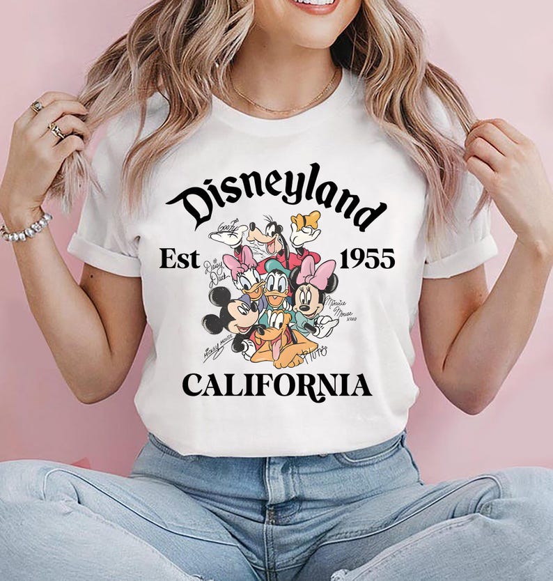 May include: White t-shirt featuring the Disneyland logo with the text "Est 1955 CALIFORNIA" in black script. The shirt showcases a colorful graphic of Disney characters including Mickey Mouse, Minnie Mouse, and Donald Duck.
