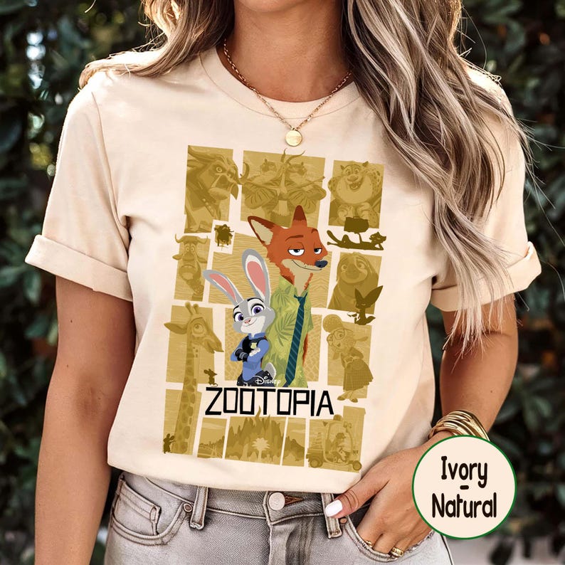 May include: Ivory-colored t-shirt featuring a graphic from the Disney movie Zootopia. The design includes characters Nick Wilde and Judy Hopps, with the movie title below. The shirt has short sleeves and a crew neck.