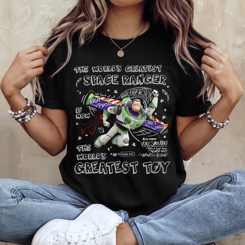 Op de afbeelding: Zwart T-shirt met Buzz Lightyear van Toy Story. De tekst "THE WORLD'S GREATEST SPACE RANGER" en "THE WORLD'S GREATEST TOY" is zichtbaar. Het ontwerp toont Buzz Lightyear in actie. Het T-shirt wordt door een persoon gedragen.