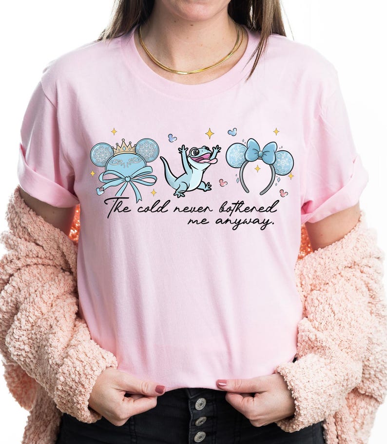 Op de afbeelding: Lichtroze T-shirt met een afbeelding van drie cartoon elementen: een Mickey Mouse oor hoofdband met een kroon, een hagedis en een Minnie Mouse oor hoofdband met een strik. De tekst "The cold never bothered me anyway" staat eronder.