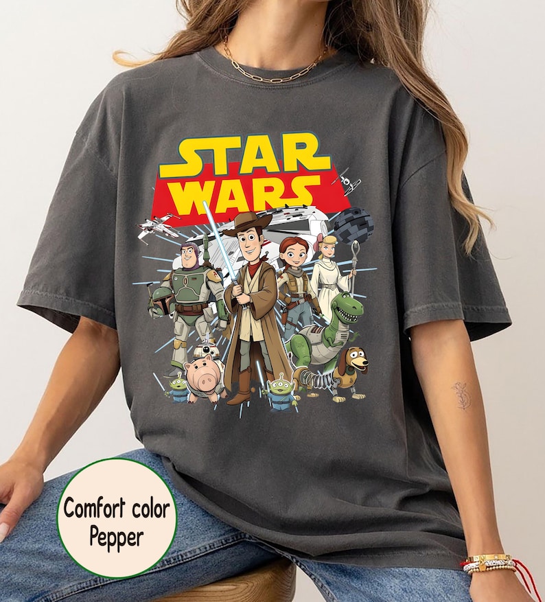 May include: Dark grey Comfort Colors t-shirt with a cartoon graphic of Star Wars characters. The Star Wars logo is in yellow and red. The t-shirt has a relaxed fit.