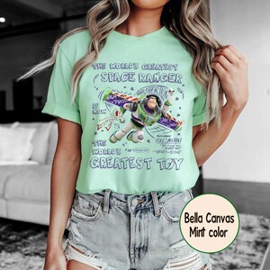 Op de afbeelding: Mintgroen t-shirt met een afbeelding van Buzz Lightyear en de tekst "THE WORLD'S GREATEST SPACE RANGER" en "THE WORLD'S GREATEST TOY". Het shirt is gemaakt van Bella Canvas materiaal.