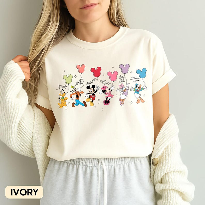 K&ouml;nnte beinhalten: Elfenbeinfarbenes T-Shirt mit einem farbenfrohen Aufdruck von Disney-Figuren, die Mickey Mouse-f&ouml;rmige Ballons halten. Die Figuren sind Pluto, Goofy, Mickey Mouse, Minnie Mouse, Daisy Duck und Donald Duck. Das Wort "Ivory" ist unten aufgedruckt.