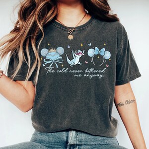 Op de afbeelding: Donkergrijs T-shirt met de tekst "The cold never bothered me anyway". Het ontwerp bevat cartoonafbeeldingen van een kroon, een hagedis en muizenoren met strikken. Het T-shirt is gemaakt van zacht materiaal.