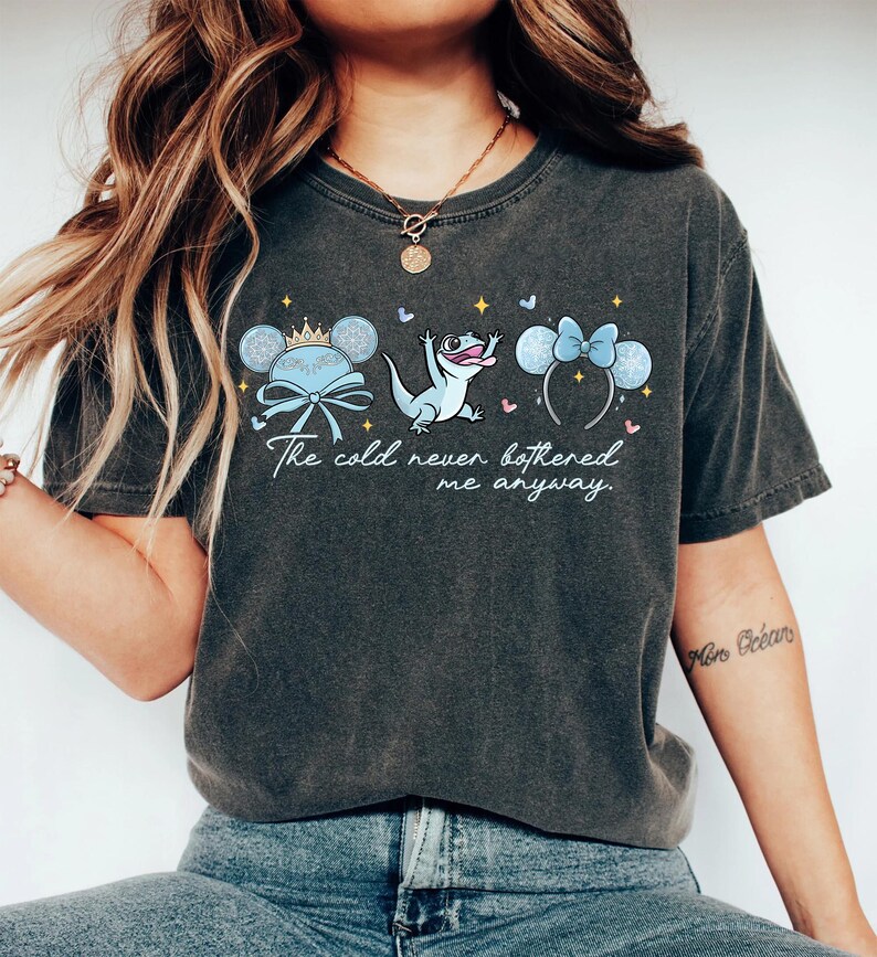 Op de afbeelding: Donkergrijs T-shirt met de tekst "The cold never bothered me anyway". Het ontwerp bevat cartoonafbeeldingen van een kroon, een hagedis en muizenoren met strikken. Het T-shirt is gemaakt van zacht materiaal.