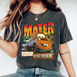 Puede incluir: Camiseta gris oscuro con una imagen de dibujos animados de Mater de la pel&iacute;cula Cars. El dise&ntilde;o incluye la palabra "MATER" en letras grandes y coloridas, y la frase "JUST KEEP TRUCKIN'". La camiseta tambi&eacute;n tiene un logotipo de salvamento de remolque.