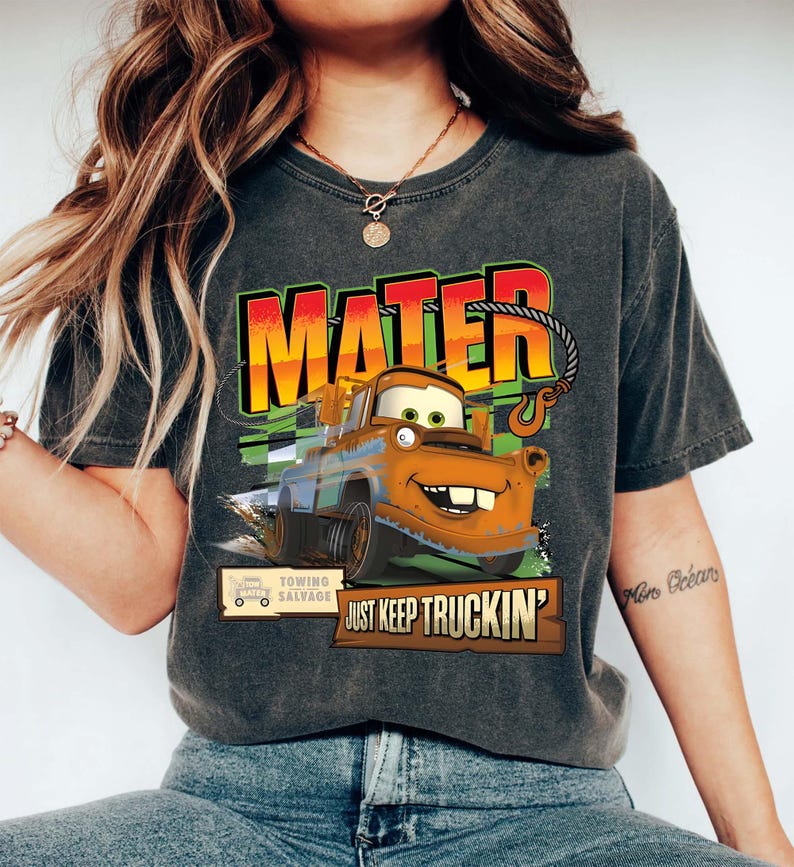 Puede incluir: Camiseta gris oscuro con una imagen de dibujos animados de Mater de la pel&iacute;cula Cars. El dise&ntilde;o incluye la palabra "MATER" en letras grandes y coloridas, y la frase "JUST KEEP TRUCKIN'". La camiseta tambi&eacute;n tiene un logotipo de salvamento de remolque.