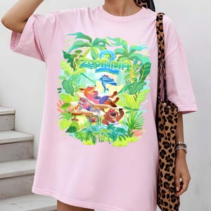 Pode incluir: Vestido camiseta rosa claro com um gr&aacute;fico colorido do filme de anima&ccedil;&atilde;o Zootopia 2. O design apresenta personagens de desenhos animados em um cen&aacute;rio tropical, com o t&iacute;tulo do filme em verde e azul. O vestido tem mangas curtas.
