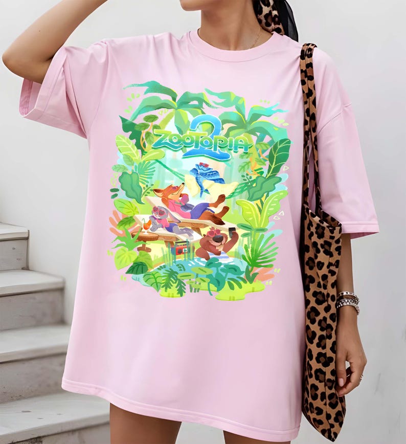 Pode incluir: Vestido camiseta rosa claro com um gr&aacute;fico colorido do filme de anima&ccedil;&atilde;o Zootopia 2. O design apresenta personagens de desenhos animados em um cen&aacute;rio tropical, com o t&iacute;tulo do filme em verde e azul. O vestido tem mangas curtas.