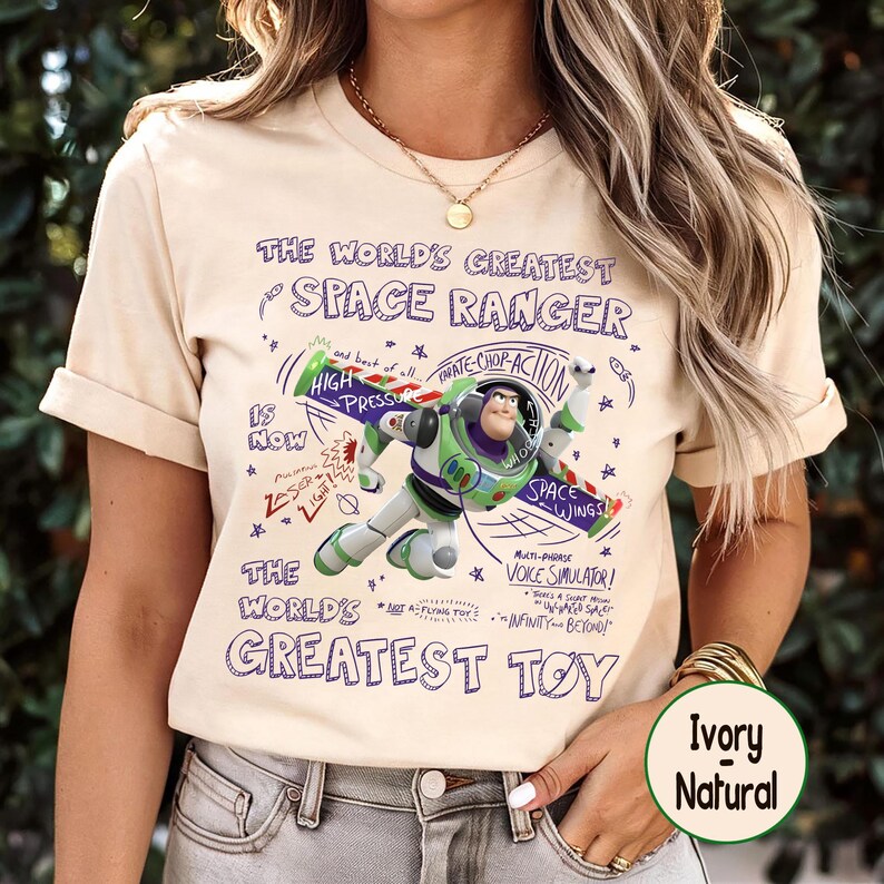 Op de afbeelding: Ivoor kleurig t-shirt met een afbeelding van Buzz Lightyear met de tekst "THE WORLD'S GREATEST SPACE RANGER" en "THE WORLD'S GREATEST TOY". Het ontwerp bevat zinnen als "High Pressure" en "Space Wings".