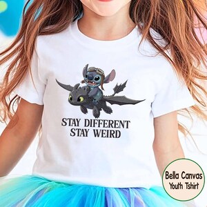 May include: White youth t-shirt with a graphic of Stitch riding Toothless, with the text "Stay Different Stay Weird". The design features a blue Stitch wearing goggles and a black dragon. Made by Bella Canvas.