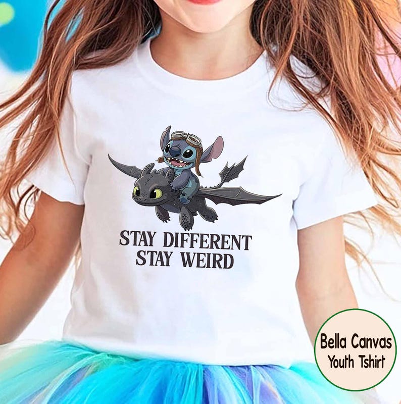 May include: White youth t-shirt with a graphic of Stitch riding Toothless, with the text "Stay Different Stay Weird". The design features a blue Stitch wearing goggles and a black dragon. Made by Bella Canvas.