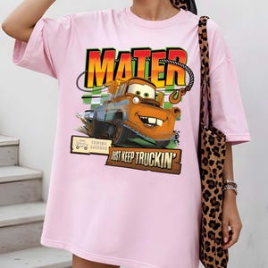 Puede incluir: Camiseta rosa claro con un gr&aacute;fico de Mater de la pel&iacute;cula Cars. El gr&aacute;fico incluye la palabra "MATER" en una fuente colorida, la imagen de Mater y la frase "JUST KEEP TRUCKIN'". La camiseta tambi&eacute;n tiene un letrero de "TOWING SALVAGE".