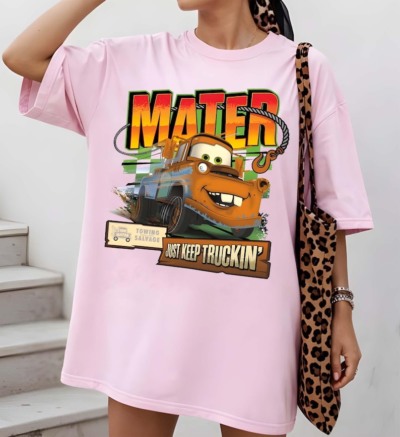 Puede incluir: Camiseta rosa claro con un gr&aacute;fico de Mater de la pel&iacute;cula Cars. El gr&aacute;fico incluye la palabra "MATER" en una fuente colorida, la imagen de Mater y la frase "JUST KEEP TRUCKIN'". La camiseta tambi&eacute;n tiene un letrero de "TOWING SALVAGE".