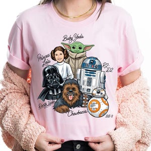 May include: Pink t-shirt featuring a Star Wars graphic with characters including Darth Vader, Princess Leia, Baby Yoda, Chewbacca, R2-D2, and BB-8. The names of the characters are written in a script font. The shirt is a crew neck with short sleeves.