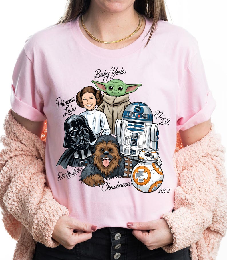 May include: Pink t-shirt featuring a Star Wars graphic with characters including Darth Vader, Princess Leia, Baby Yoda, Chewbacca, R2-D2, and BB-8. The names of the characters are written in a script font. The shirt is a crew neck with short sleeves.