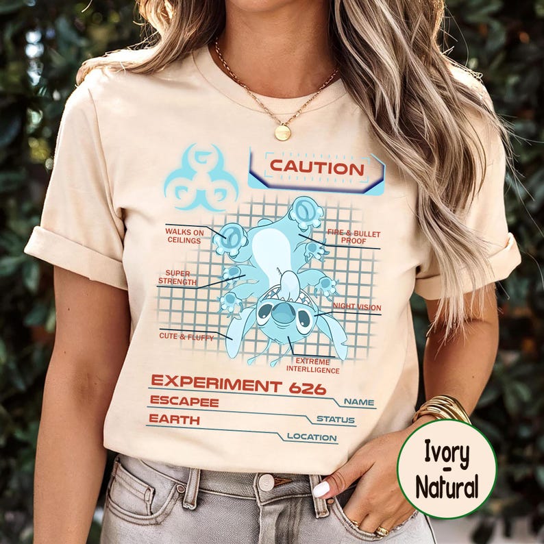 May include: Ivory-colored t-shirt featuring a light blue cartoon character illustration with the text "CAUTION" and "EXPERIMENT 626." The design includes the character's abilities: walks on ceilings, super strength, and night vision. The shirt is made of a soft material.