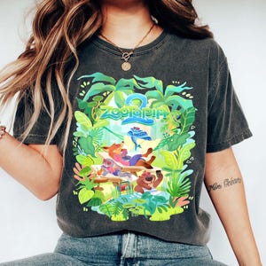 Pode incluir: T-shirt cinzento escuro com um gr&aacute;fico colorido de "Zootopia 2". O design apresenta o t&iacute;tulo do filme e personagens de desenhos animados relaxando em um cen&aacute;rio tropical, rodeados por folhagens verdes.