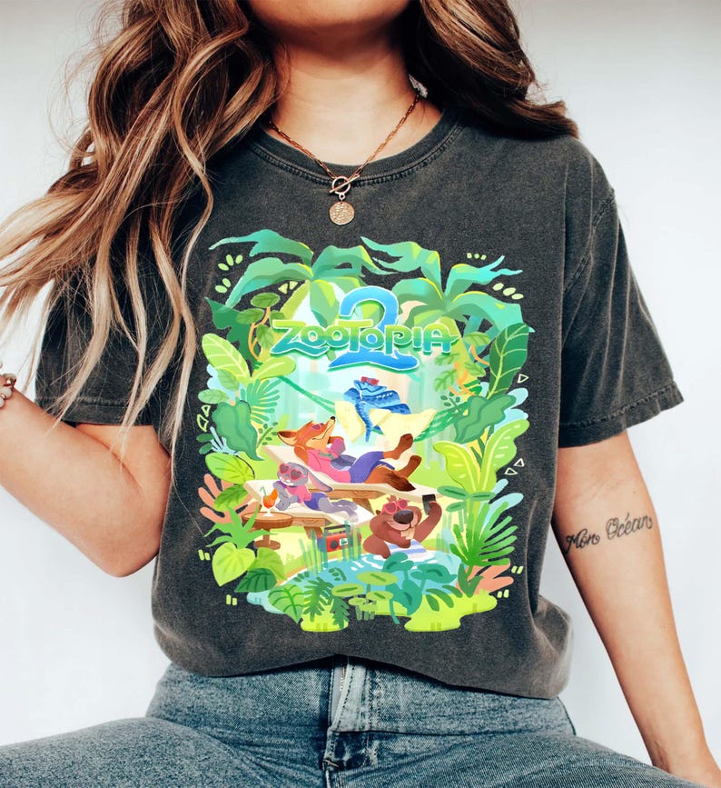 Pode incluir: T-shirt cinzento escuro com um gr&aacute;fico colorido de "Zootopia 2". O design apresenta o t&iacute;tulo do filme e personagens de desenhos animados relaxando em um cen&aacute;rio tropical, rodeados por folhagens verdes.