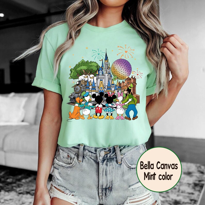 Peut inclure: T-shirt vert menthe avec un motif color&eacute; de personnages Disney devant un ch&acirc;teau, une grande sph&egrave;re et un train. Le texte "Bella Canvas Mint color" est dans un cercle en bas &agrave; droite.