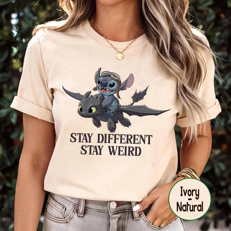 May include: Cream-coloured t-shirt with a cartoon image of Stitch riding a dragon, and the words "STAY DIFFERENT STAY WEIRD". The design is printed in black. The shirt has short sleeves and is made of a soft material.