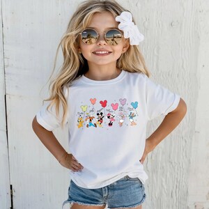 K&ouml;nnte beinhalten: Wei&szlig;es T-Shirt mit einem farbenfrohen Aufdruck von Disney-Figuren, die Ballonstr&auml;u&szlig;e halten. Die Figuren sind Pluto, Goofy, Mickey Mouse, Minnie Mouse, Daisy Duck und Donald Duck. Die Ballons sind in verschiedenen Farben.