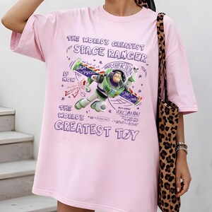 Op de afbeelding: Een lichtroze t-shirt met een afbeelding van Buzz Lightyear met de tekst "THE WORLD'S GREATEST SPACE RANGER" en "THE WORLD'S GREATEST TOY". Het ontwerp bevat zinnen als "High Pressure" en "Space Wings".