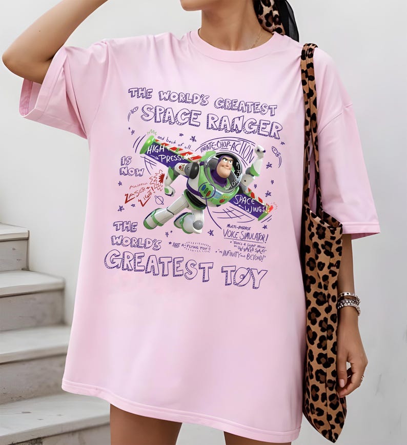 Op de afbeelding: Een lichtroze t-shirt met een afbeelding van Buzz Lightyear met de tekst "THE WORLD'S GREATEST SPACE RANGER" en "THE WORLD'S GREATEST TOY". Het ontwerp bevat zinnen als "High Pressure" en "Space Wings".